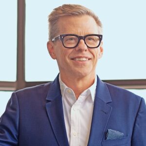 Gerry Zack; smiling white man wearing a dark rimmed glasses, white collared shirt, no tie, and abright blue blazer