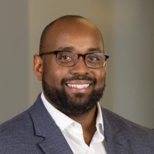 Image of Brian Stanley; smiling African American man wearing glasses, a white collared shirt, no tie, and a grey blazer.